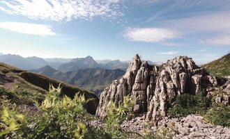 An impressive mountain landscape with rugged rocks and majestic mountains in the background. The sky is clear and the surroundings are characterized by green vegetation. | © Kleinwalsertal Tourismus eGen | Frank Drechsel