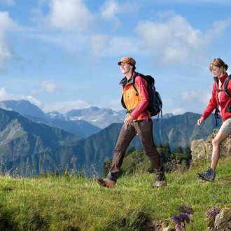 © Kleinwalsertal Tourismus | Andi Mayr Two hikers are walking through a green landscape with mountains in the background. The sky is blue with some clouds. | © Kleinwalsertal Tourismus | Andi Mayr
