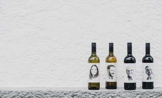 Four bottles of wine are placed on a gray surface. The labels display black-and-white portraits of different individuals. | © Das Naturhotel Chesa Valisa
