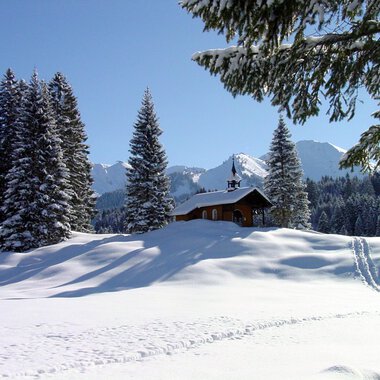 A snowy landscape with tall fir trees and a small wooden house. In the background, the snow-covered mountains and a clear blue sky can be seen. | © Bruder Klaus Kapelle | Rolf Koeberle