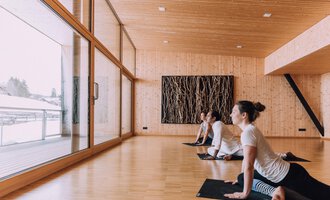 A bright yoga room with wooden paneling and large windows. Three people are practicing yoga and looking outside. | © Naturhotel Chesa Valisa