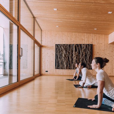 A bright yoga room with wooden paneling and large windows. Three people are practicing yoga and looking outside. | © Naturhotel Chesa Valisa