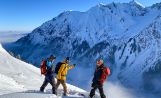 Three people in winter gear are standing on a snow-covered mountain. In the background, impressive mountains stretch under a clear blue sky.