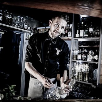 Ein Barkeeper, der an einer gut gefüllten Bar arbeitet. Er lächelt, während er mit einem Glas beschäftigt ist. | © Verwöhn- und Wellnesshotel Walserhof | Werner Krug