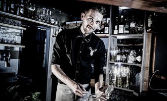 Ein Barkeeper, der an einer gut gefüllten Bar arbeitet. Er lächelt, während er mit einem Glas beschäftigt ist. | © Verwöhn- und Wellnesshotel Walserhof | Werner Krug
