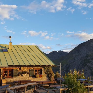 A cozy mountain hut with a green roof in the Alps. In the background, majestic mountains stretch out under a blue sky. | © Alpe Kuhgehren | Lisa Hiesinger