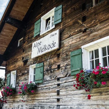 A traditional wooden house with green shutters. Flower boxes with colorful flowers adorn the windows. | © Kleinwalsertal Tourismus | Andre Tappe