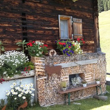 A traditional wooden house with colorful flowers and wood piles. The place radiates rural coziness and a connection to nature. | © Kleinwalsertal Tourismus | N. Lughammer