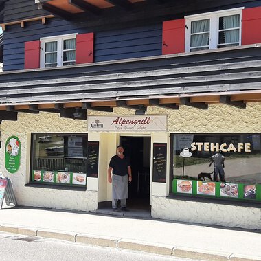 A traditional restaurant with an inviting entrance and a terrace. The facade is designed in light and dark colors, with a small group of menus and offers on posters. | © Alpengrill Riezlern | Veronika Senn