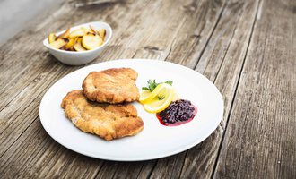 A plate with two breaded schnitzels, served with lemon slices and a portion of cranberry sauce. In the background, there is a bowl of fries. | © Alpenhotel DAS KÜREN
