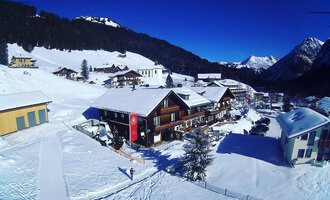 A snow-covered landscape with a cozy wooden house and mountains in the background. The sky is clear and blue.