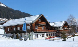 A traditional wooden house in the snow, surrounded by mountains. The sky is clear and blue, and there are many snow-covered areas.