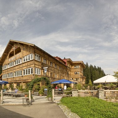 A rustic hotel with a wooden facade in a beautiful natural landscape. The outdoor area has seating and sun umbrellas under a cloudy sky. | © Auenhütte | Yorck Dertinger