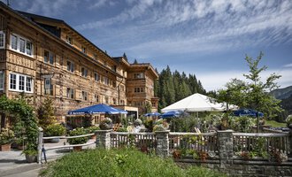 A traditional wooden building in a picturesque setting. In front of the building, there is a beautiful garden and a tent with blue umbrellas. | © Auenhütte | Yorck Dertinger