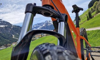An orange mountain bike is standing on a green hill overlooking the mountains. The sky is clear and it is a sunny day. | © Austrian Ski- & Service Ranch | Trixi Leitner