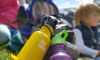 Two colorful water bottles in yellow and purple are in the foreground on a green meadow. In the background, children and a backpack can be seen, with mountains and a blue sky. | © Austrian Ski- & Service Ranch | Trixi Leitner