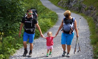 A family is hiking on a narrow path in nature. The parents are holding their little daughter's hands and carrying backpacks. | © Austrian Ski- & Service Ranch | Trixi Leitner