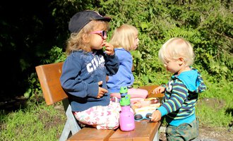 Three children are sitting on a bench outside. They are enjoying snacks and playing together. | © Kleinwalsertal Tourismus | Britta Maier