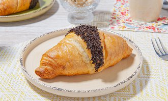A delicious croissant with chocolate glaze is lying on a light plate. In the background, more breakfast items are visible. | © MPREIS Warenvertriebs GmbH
