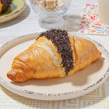 A delicious croissant with chocolate glaze is lying on a light plate. In the background, more breakfast items are visible. | © MPREIS Warenvertriebs GmbH