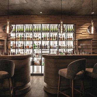 A stylish bar with wooden interiors and a lit shelf full of bottles. The seating is cozy and modern. | © Travel Charme Ifen Hotel