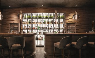 A stylish bar with wooden interiors and a lit shelf full of bottles. The seating is cozy and modern. | © Travel Charme Ifen Hotel