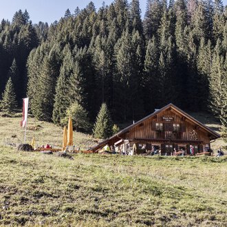 A cozy mountain cabin surrounded by dense, green forests. In the foreground, there is a meadow with colorful flags. | © Kleinwalsertal Tourismus | Bastian Morell