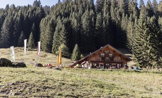 A cozy mountain cabin surrounded by dense, green forests. In the foreground, there is a meadow with colorful flags. | © Kleinwalsertal Tourismus | Bastian Morell