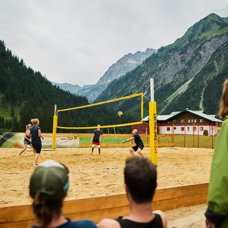 A beach volleyball game in the mountains, surrounded by a picturesque landscape. The players are actively on the sand court while spectators watch. | © Kleinwalsertal Tourismus | Oliver Farys