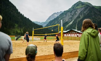 A beach volleyball game in the mountains, surrounded by a picturesque landscape. The players are actively on the sand court while spectators watch. | © Kleinwalsertal Tourismus | Oliver Farys