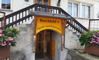 An entrance area to Berchtold's bacon and cheese cellar. Above the door hangs a yellow sign, surrounded by flowers. | © Kleinwalsertal Tourismus | N. Lughammer