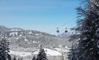 A snowy landscape with snow-covered trees and mountains in the background. Two gondolas float over the winter landscape under a clear blue sky. | © OBERSTDORF · KLEINWALSERTAL BERGBAHNEN