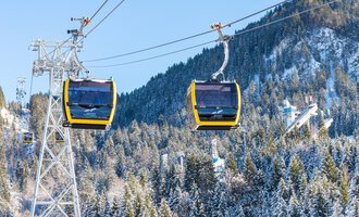 A cable car with yellow gondolas glides over snow-covered mountains and forests. The sky is clear and blue. | © OBERSTDORF · KLEINWALSERTAL BERGBAHNEN