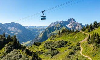 A cable car glides over green hills and mountains. The sky is clear and the landscape is picturesque. | © OBERSTDORF · KLEINWALSERTAL BERGBAHNEN