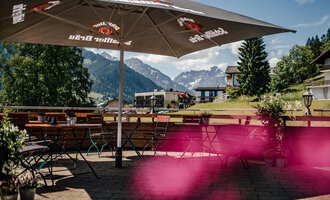 A cozy terrace with a large sun umbrella and a view of the mountains. Green trees and an inviting environment create a relaxed atmosphere.
