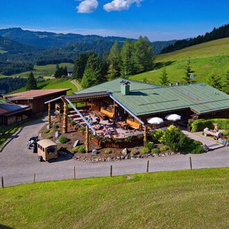 A picturesque mountain hut surrounded by green meadows and woods. On the terrace, people enjoy the sunny view of the mountains. | © Bergstüble | Jürgen Waffenschmidt