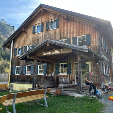 A rustic wooden house named "Gemstelhof" with an inviting porch. In the foreground, you can see a person sitting on a bench, surrounded by green landscape and mountains. | © Bernhards Gemstel Hofladen | Bernhard Heim