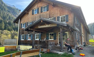 A traditional wooden house named "Gemstelhof" stands in a rural setting. Benches are placed in front of the building, and a person is visible, while the mountains are seen in the background. | © Bernhards Gemstel Hofladen | Bernhard Heim