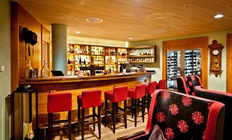 A cozy bar with a long wooden counter and red bar stools. In the background, shelves with drinks and a modern clock are visible. | © Hotel Birkenhöhe | Fotograf Krug