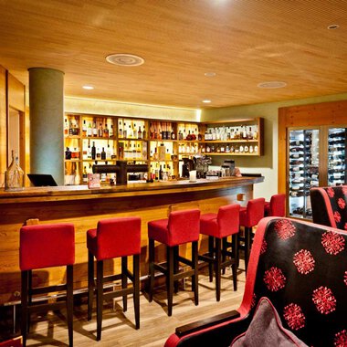 A cozy bar with a long wooden counter and red bar stools. In the background, shelves with drinks and a modern clock are visible. | © Hotel Birkenhöhe | Fotograf Krug