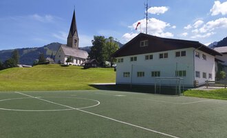 A football pitch in front of a modern building with a church in the background. The surroundings are green with hills and a clear blue sky. | © Kleinwalsertal Tourismus | N. Lughammer