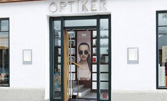 An optical shop with large windows and a distinctive advertising figure. The entrance door is open and invites customers in. | © Bräuer Optik & Akustik | AlpenMedia-Design