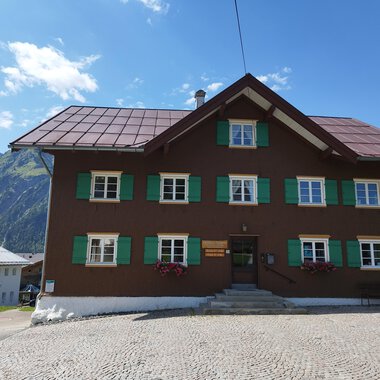 Ein traditionelles altes Haus mit brauner Fassade und grünen Fensterläden. Im Hintergrund sind Berge und ein blauer Himmel sichtbar. | © Kleinwalsertal Tourismus | Nicole Lughammer