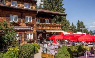 A charming inn with a wooden facade and blooming flowers. Outside, tables are set up with red umbrellas. | © Bühlalpe | Frank Drechsel