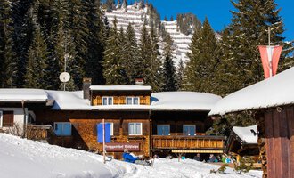 A cozy mountain cabin in winter with a snow-covered ground. In the background, high mountains and green fir trees can be seen. | © Bühlalpe | Frank Drechsel