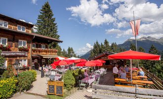 A cozy restaurant with a terrace, surrounded by nature and mountains. Guests sit under red umbrellas and enjoy the view. | © Bühlalpe | Frank Drechsel