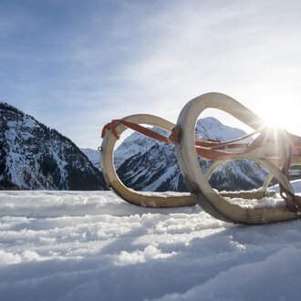 A sled lies in the snow, surrounded by a wintry mountain landscape. The sun shines out from behind the mountains. | © Kleinwalsertal Tourismus | Dominik Berchtold