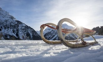 A sled lies in the snow, surrounded by mountains. The sun shines in the background, creating a wintry atmosphere. | © Kleinwalsertal Tourismus | Dominik Berchtold