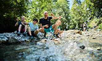 A group of children is sitting by the shore of a crystal-clear stream and having fun. In the background, green trees and a summery landscape can be seen. | © Kleinwalsertal Tourismus | Oliver Farys