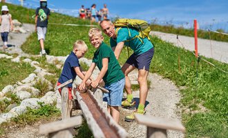Father and his two sons are playing on a path outdoors. In the background, more people can be seen, and the surroundings are green and sunny. | © OBERSTDORF · KLEINWALSERTAL BERGBAHNEN